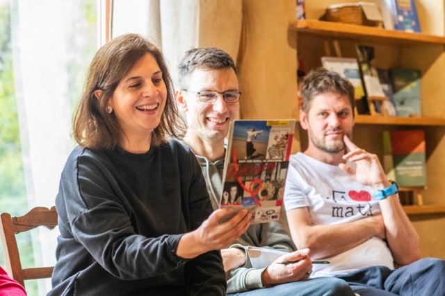 Účastníci sedia v interiéri počas workshopu, žena ukazuje kartičku a ostatní sa usmievajú.